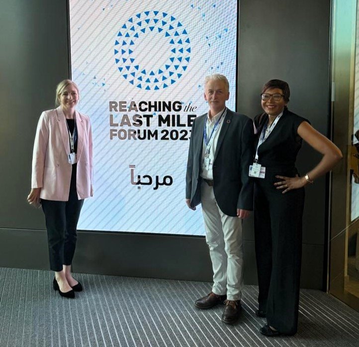 Uniting to Combat NTDs staff Louisa Tribe and Stuart Halford and Board Member Dr Francisca Mutapi in front of RLMF signage